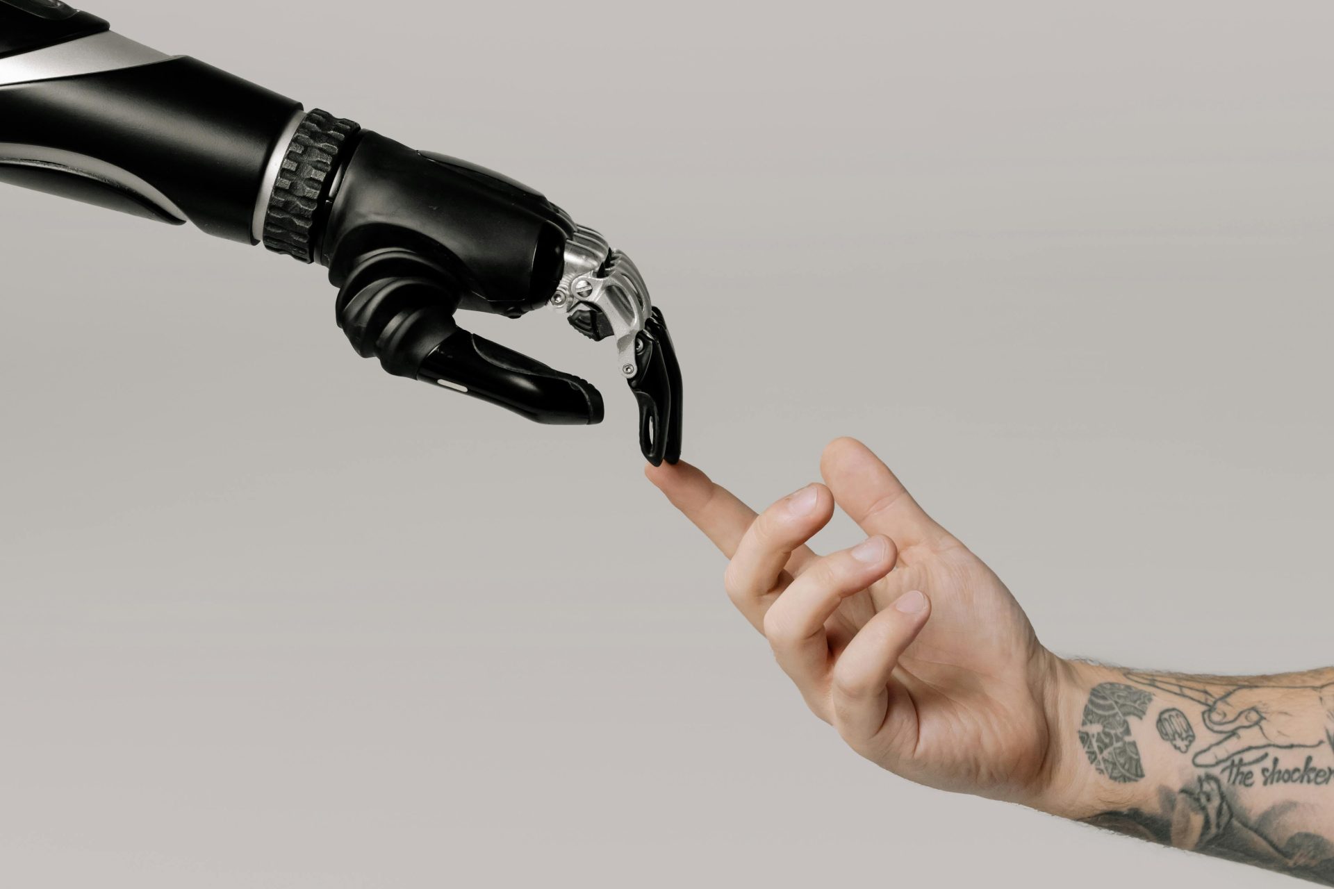 Eine menschliche Hand mit Tätowierungen streckt sich auf weißem Hintergrund nach einer Roboterhand aus.